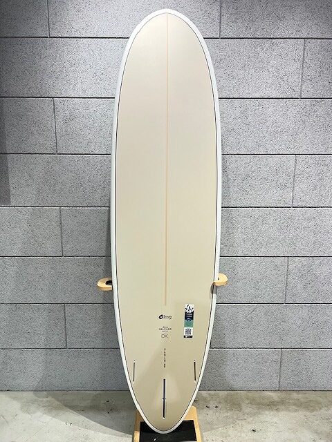 Cream-colored longboard surfboard with a subtle orange stringer, standing on a wooden rack against a gray block wall.