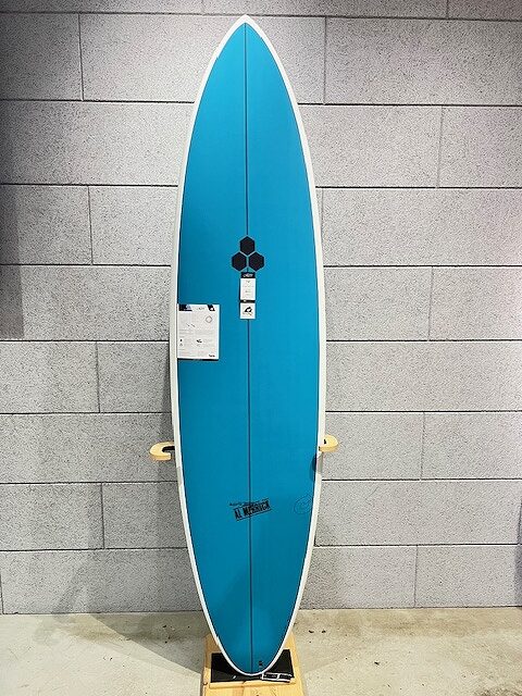 Blue surfboard on a display stand, standing upright against a gray brick wall with tags attached.