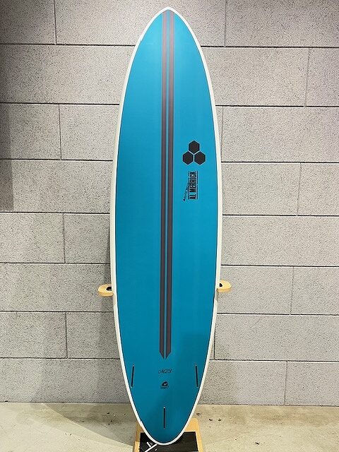 Blue surfboard standing upright against a gray brick wall on a rack, showing a central dark stripe and small logos near the top.