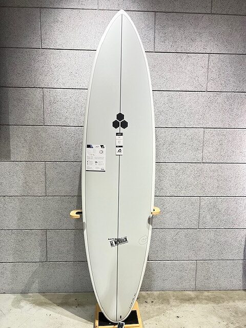 White surfboard standing upright on a wooden rack against a gray tiled wall, with stickers and a black hexagon logo in the center.