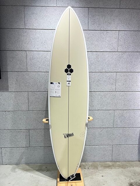 Light yellow surfboard standing upright against a gray brick wall on a wooden stand, with stickers and a hexagon logo near the center tail area.