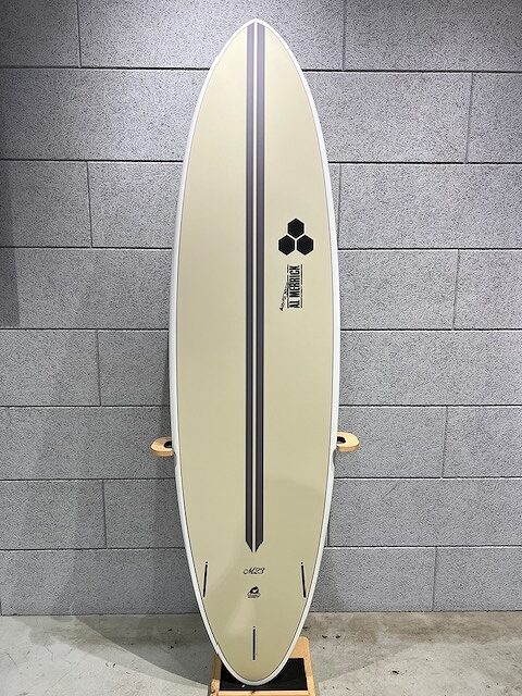 Cream-colored surfboard with a double dark center stripe, standing upright against a gray brick wall with a hexagon logo near the top.