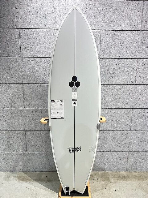 White-gray surfboard standing upright against a gray wall, with a black hexagon logo and several tags visible on the board.
