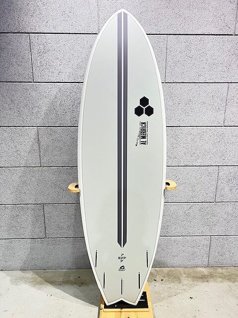 Light gray surfboard with a double dark blue center stripe, standing upright on a stand against a gray brick wall; features a hexagon logo and 'Al Merrick' text along the stripe