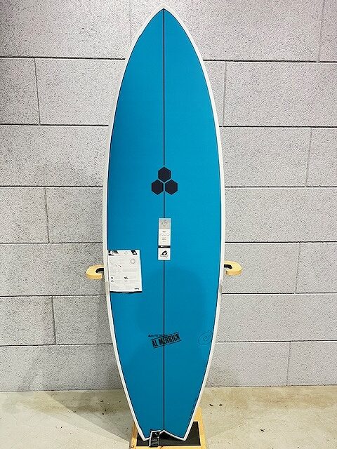 Blue surfboard with a white edge standing upright against a gray brick wall, with product tags attached to it.