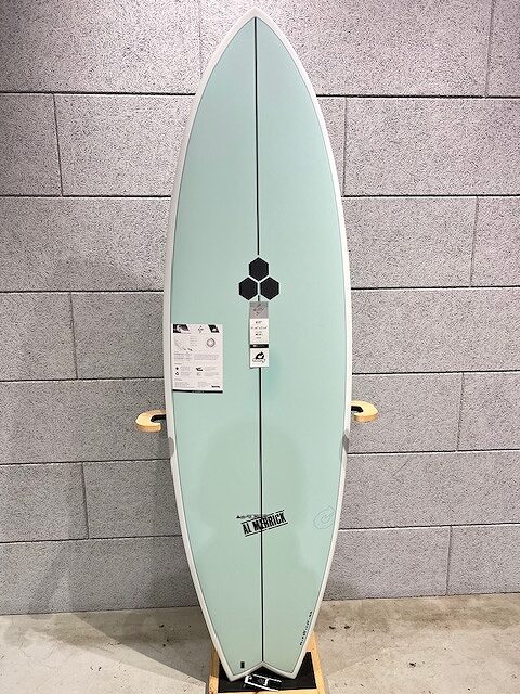 Mint-green surfboard standing upright on a wooden rack against a gray tiled wall in a shop. The board has white rails and a few labels attached near center.