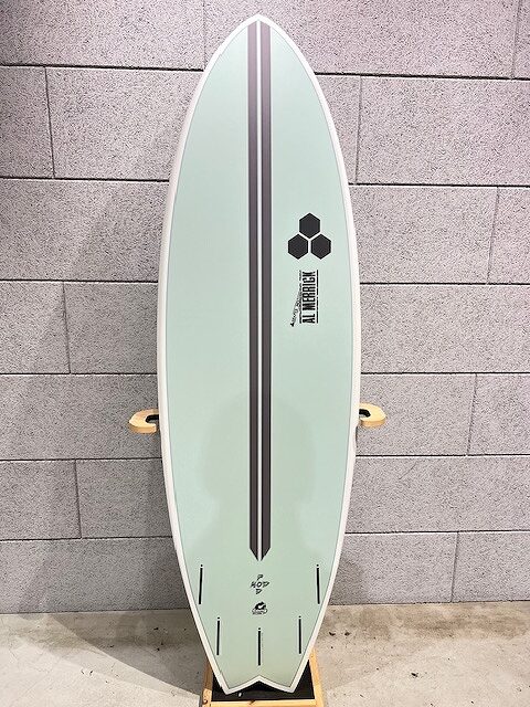 Mint green surfboard with a black center stripe and logos, standing upright on a rack against a gray brick wall. No fins attached visible, just yellow bushings on the rack beside it.