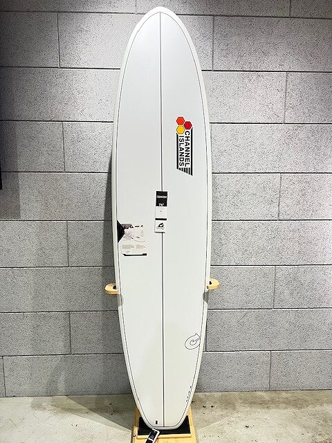 White surfboard standing upright against a gray tiled wall, featuring a Channel Islands sticker toward the top and a small black label midboard.