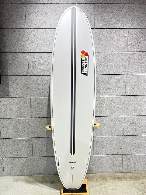 White surfboard with a dark central stripe on a stand against a gray brick wall; near the top is a Stanley Islands sticker.