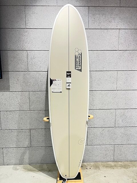 Cream-colored Channel Islands surfboard on a stand against a gray brick wall, with price/info tags attached.