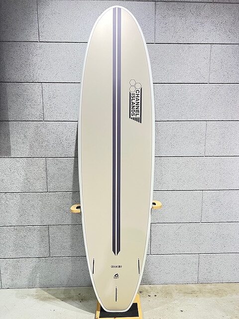 Cream-colored surfboard with a dark blue double stripe down the center, Channel Islands logo near the top, standing upright against a gray tiled wall.