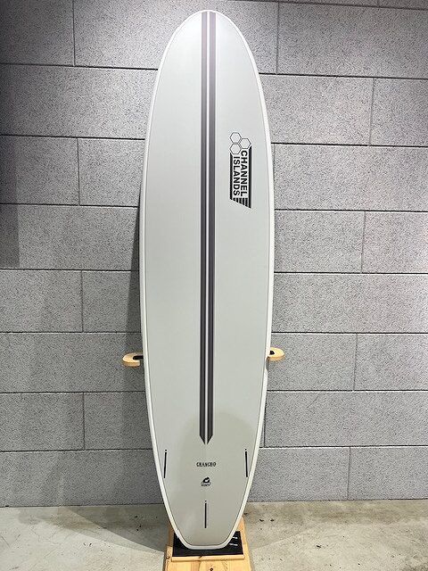 White surfboard standing upright on a wooden rack against a gray tiled wall, featuring a dark center stringer and a Channel Islands logo toward the upper half.