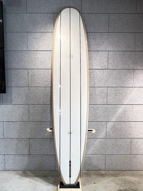 White longboard surfboard standing upright on a display stand against a gray brick wall interior.