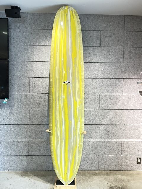 s-IMG_5672 - SHOP NEWS | ムラサキスポーツオンラインストア 通販 Yellow and clear striped surfboard wrapped in plastic, standing upright against a gray tiled wall indoors.