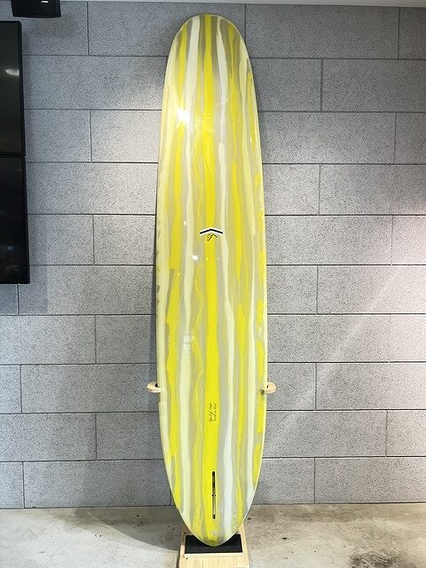 s-IMG_5673 - SHOP NEWS | ムラサキスポーツオンラインストア 通販 Surfboard with yellow and gray vertical stripes standing upright on a wooden stand against a gray tiled wall.