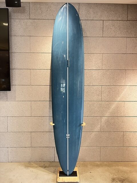 Blue surfboard standing upright on a wooden display stand against a gray brick wall.