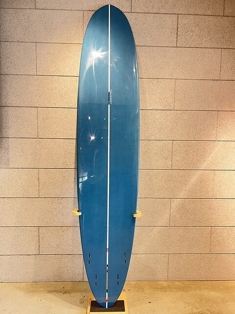 Blue surfboard standing upright against a beige brick wall on a wooden stand indoors, with a white center seam running lengthwise.