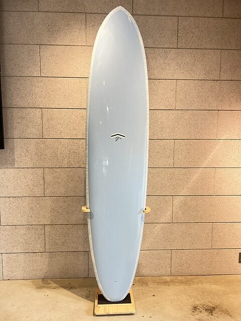 s-IMG_5678 - SHOP NEWS | ムラサキスポーツオンラインストア 通販 Light blue surfboard standing upright on a wooden rack indoors against a beige brick wall.