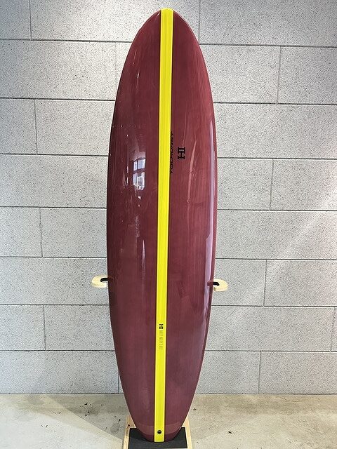 Maroon surfboard with a bright yellow center stripe standing upright against a gray tile wall, indoors on a display stand.