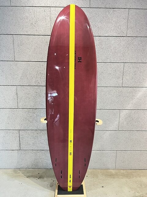 Maroon surfboard with a bright yellow center stripe standing upright against a gray brick wall.