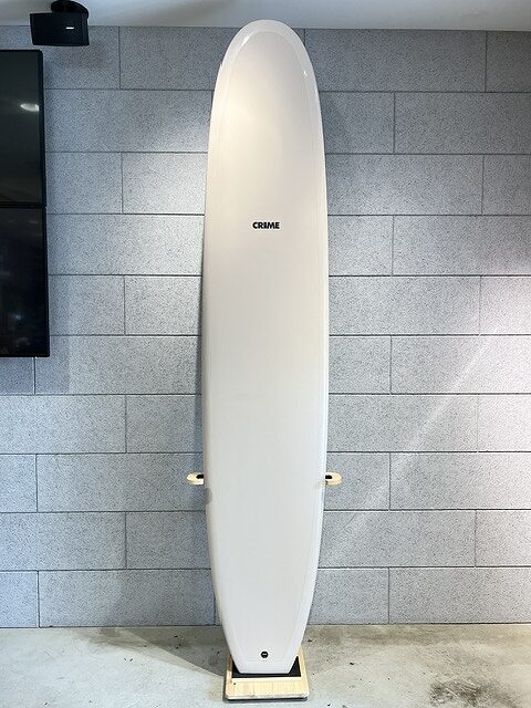 White surfboard standing upright on a wooden stand against a gray brick wall, ready for display or storage.