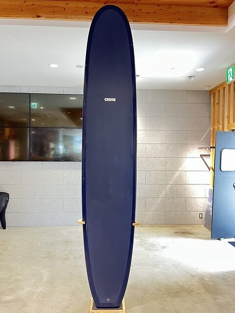 Tall blue surfboard standing upright on a wooden stand in an indoor room with gray tiled walls.