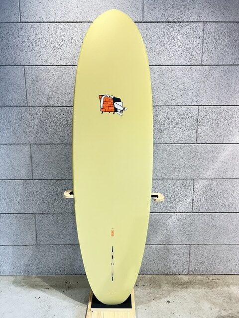 Yellow surfboard standing upright against a gray brick wall, featuring a small logo near the upper center.