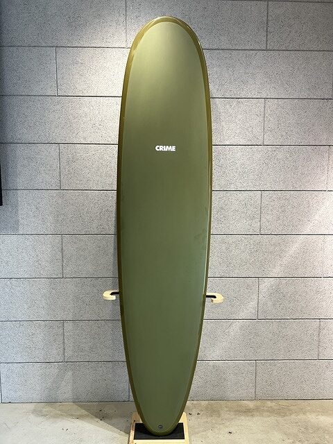 Vertical olive-green surfboard standing on a wooden stand against a gray brick wall, with the word 'CRIME' near the top.