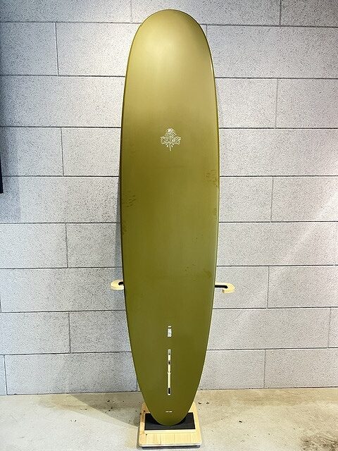 Olive-green longboard surfboard standing upright on a wooden stand against a gray brick wall, with a small logo near the center.