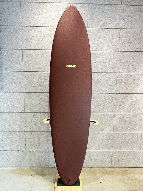 Maroon surfboard standing upright on a wooden rack against a gray brick wall, with 'CRIME' logo visible near the center.