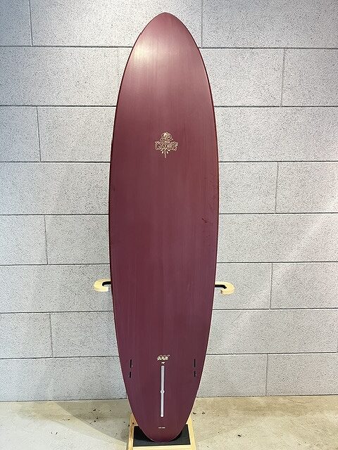Maroon longboard standing upright on a display stand against a gray tiled wall, with gold crest logos near the top and tail lengthwise.