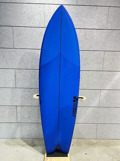 Blue surfboard standing upright on a small stand against a gray brick wall, with a center stringer and small cream-colored side fins visible.