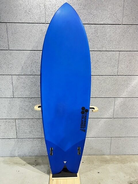 Blue surfboard standing upright against a gray brick wall, with two fin boxes near the tail.