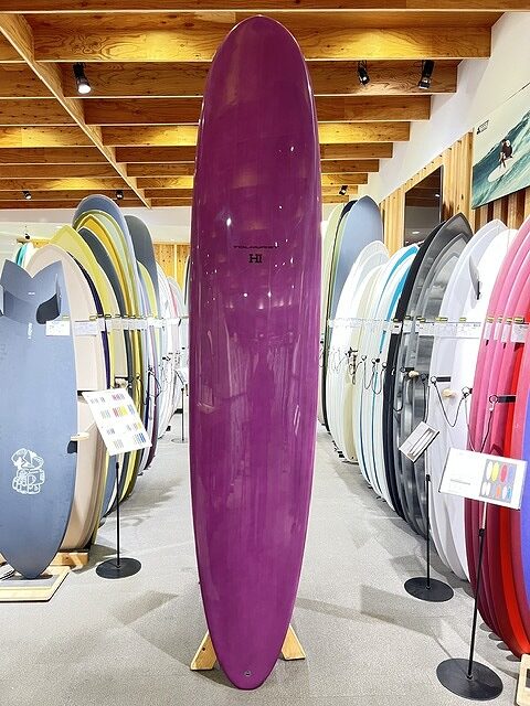 Purple surfboard standing upright on a wooden stand in a surf shop showroom, with rows of colorful boards along the walls.