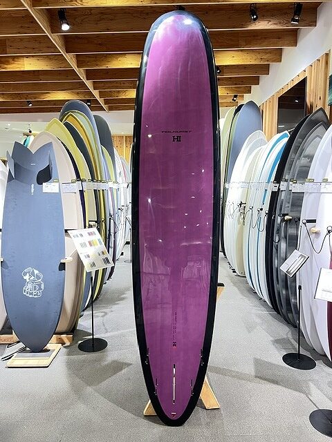 Purple longboard standing upright on a wooden rack in a surf shop, surrounded by colorful boards on both sides.