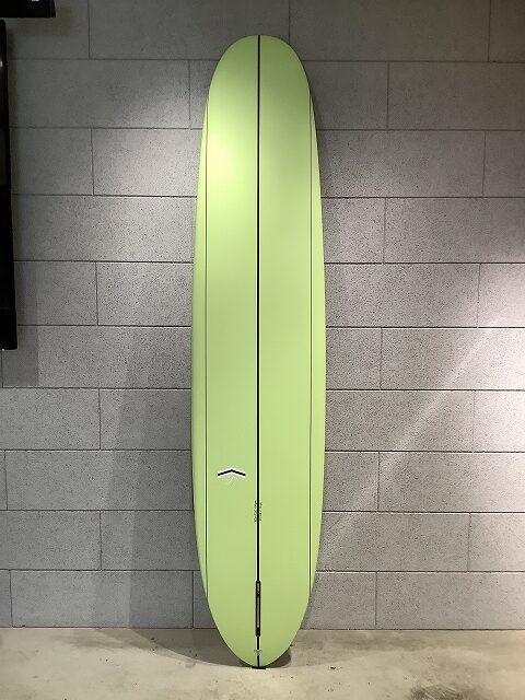 Lime-green surfboard standing upright against a gray concrete block wall, showing a long, slim shape with a center seam.