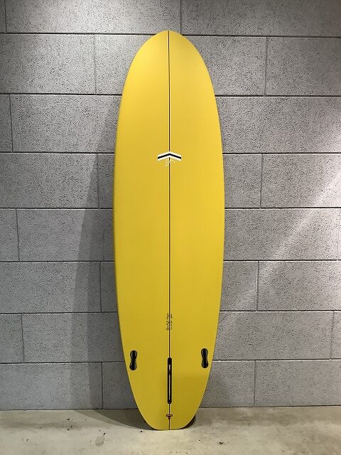 Yellow surfboard standing upright against a gray tiled wall, showing two fin boxes and a bottom leash plug near the tail.