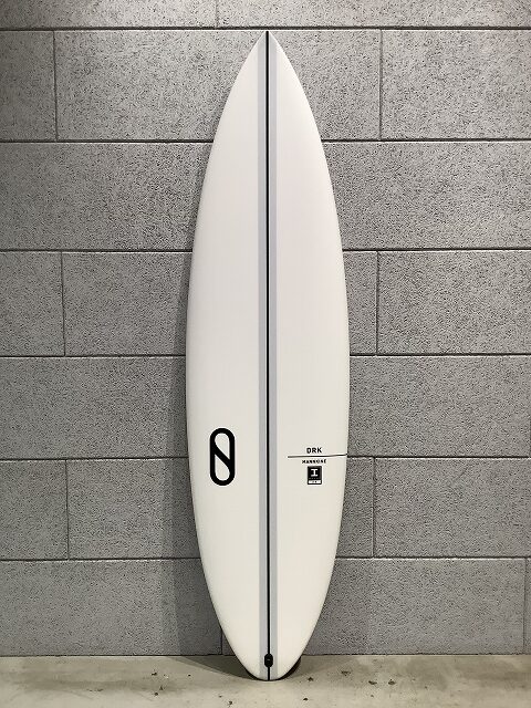 White surfboard standing upright against a gray concrete block wall, showing a dark center stringer and a black logo toward the tail.