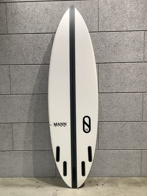 White surfboard with a black center stringer and four fin boxes, standing against a gray cinder block wall; features Mann branding and a black oval logo on the right.