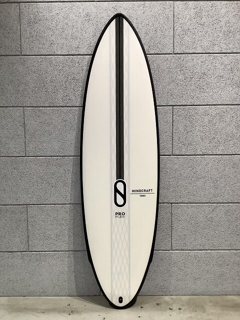 White surfboard with a black center stripe, pro model, leaning against a gray brick wall; Mindcraft Tokyo branding visible.
