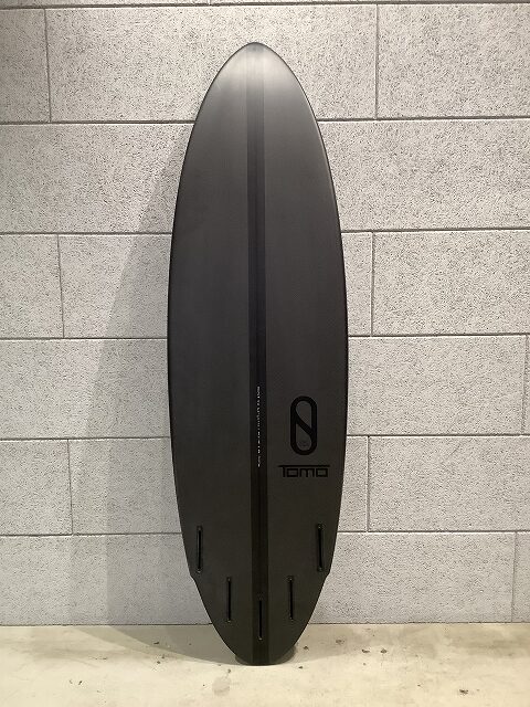 Dark gray surfboard standing upright against a light gray brick wall, with a center groove and four fin boxes at the bottom, featuring a 'TOMO' logo.