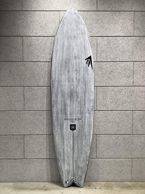 Grey surfboard with faint wood grain, standing upright against a light gray tiled wall; visible logos near the center and tail area.