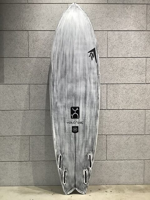 Gray surfboard standing upright against a tiled wall, featuring brand logos along its deck.