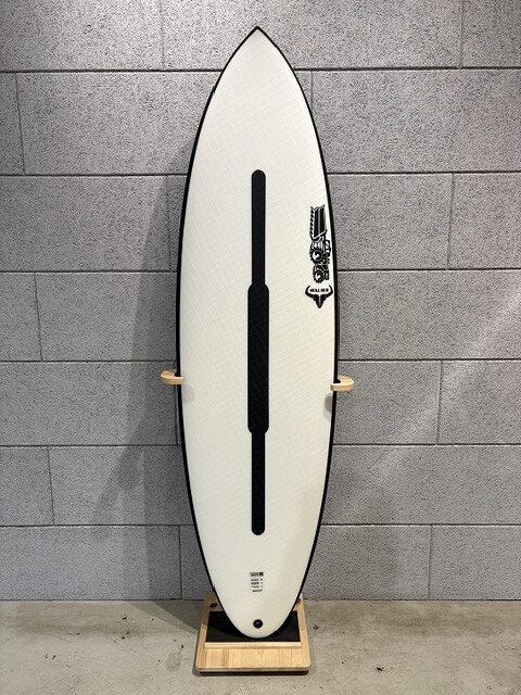 White surfboard on a wooden rack indoors, with a black center stringer and a right-side logo against a grey cinder-block wall.