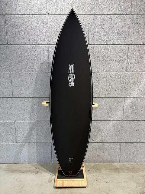 Tall black longboard standing vertically on a wooden stand against a gray cinder block wall, with a small white graphic in the center.