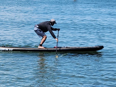 Person standing on a paddleboard, wearing a black long-sleeve shirt and striped shorts, paddling on blue water.
