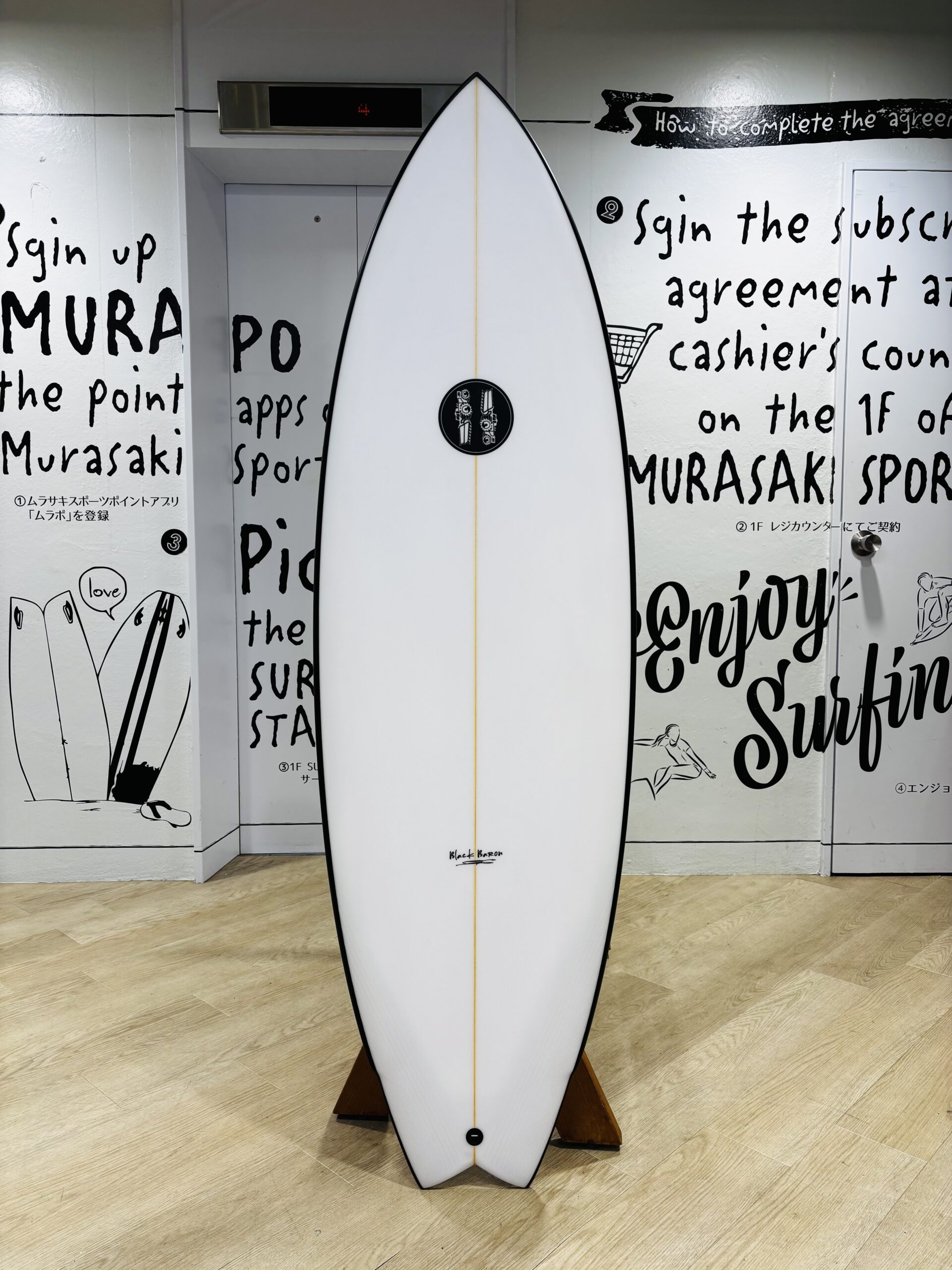 IMG_0781 - SHOP NEWS | ムラサキスポーツオンラインストア 通販 White surfboard standing upright on a wooden rack indoors, with black graffiti-style wall text in the background.