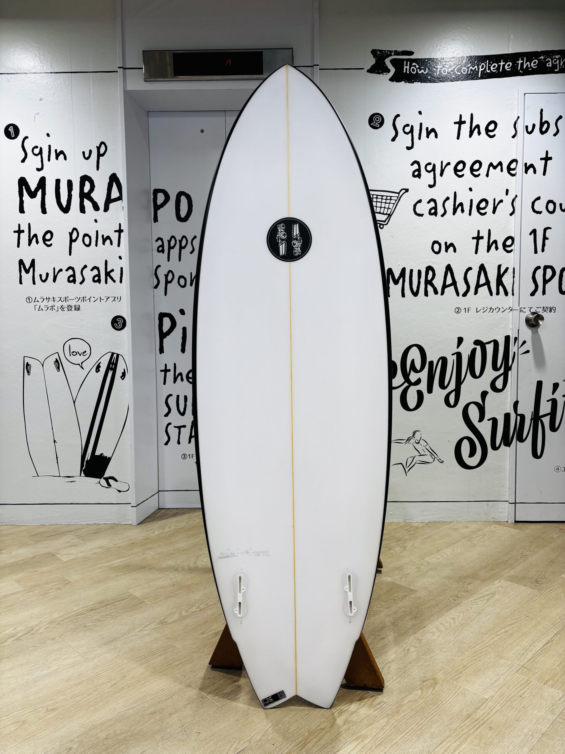IMG_0784 - SHOP NEWS | ムラサキスポーツオンラインストア 通販 White surfboard standing upright on a wooden display rack indoors, with black graffiti-style text and drawings on the walls behind it, and a small circular logo near the center.