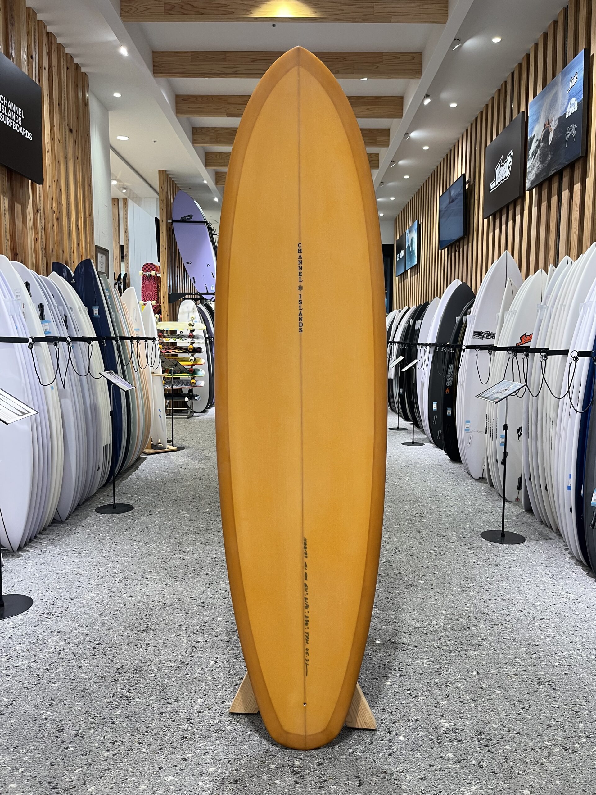 IMG_2667 - SHOP NEWS | ムラサキスポーツオンラインストア 通販 Orange surfboard standing upright in the center of a shop aisle, flanked by rows of white and colored boards on racks.