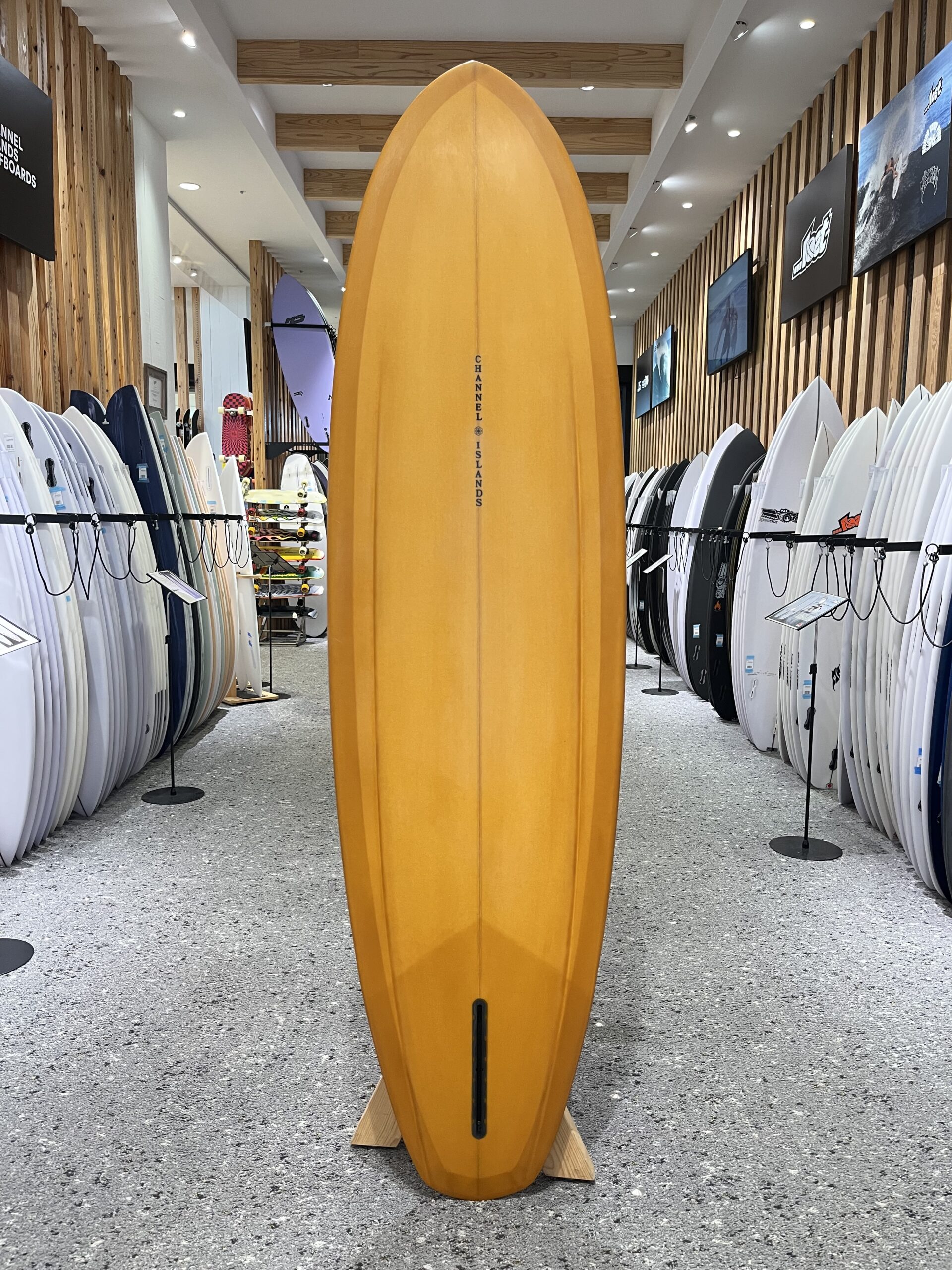 IMG_2670 - SHOP NEWS | ムラサキスポーツオンラインストア 通販 Tall orange surfboard displayed upright in a surf shop aisle, with rows of white and colored boards on both sides.